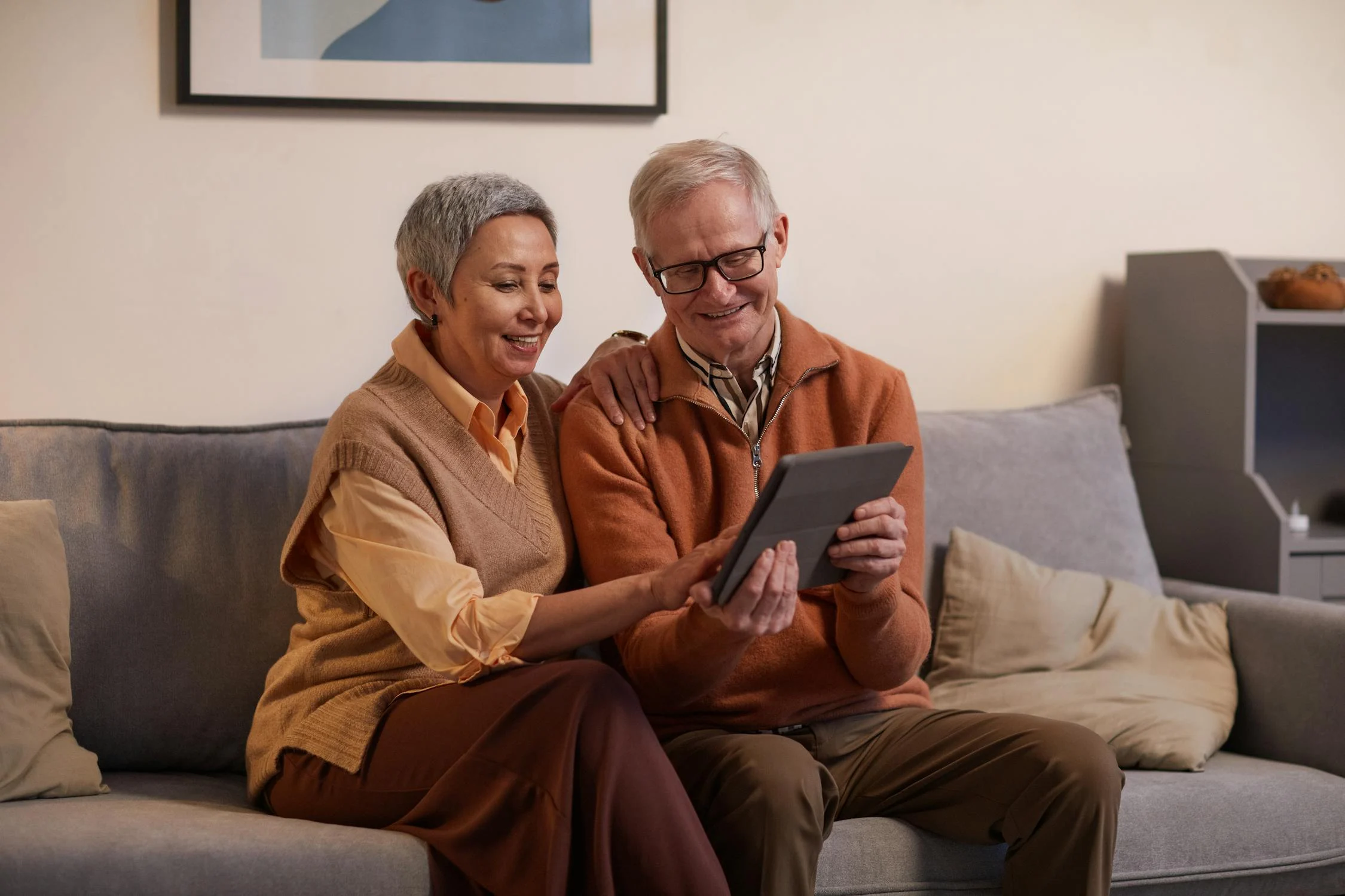 Un couple de personnes âgées, assis dans un canapé, regarde une tablette en souriant.